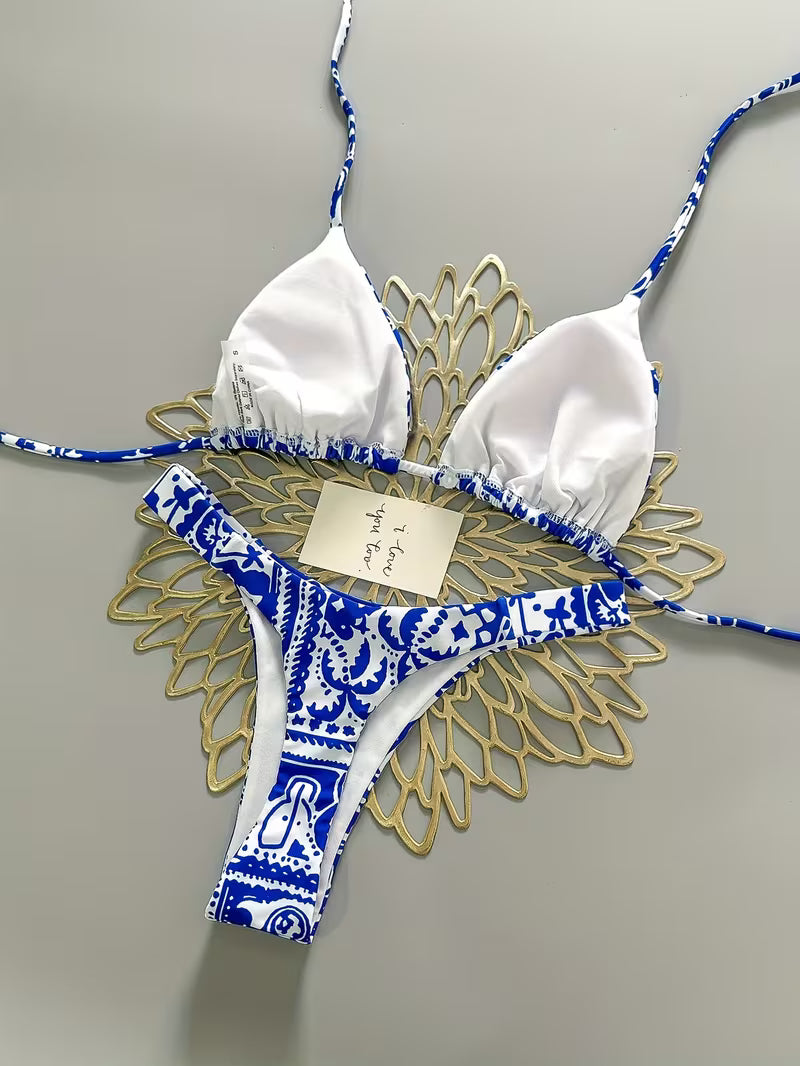 White and blue patterned string bikini laid flat on a gold decorative stand against a light gray background.