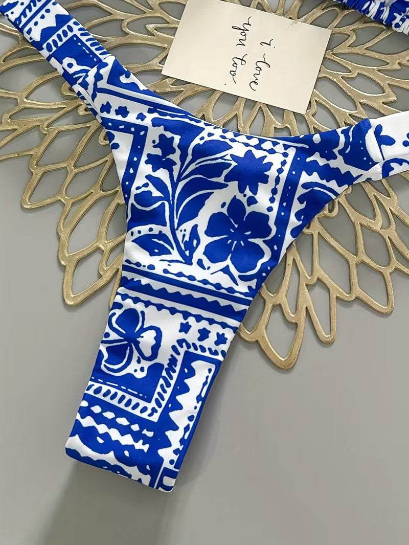 A close-up of a bikini bottom with a bold blue and white floral and geometric pattern, laid flat on a gold-colored decorative doily over a gray surface. A small handwritten note is tucked near the top.