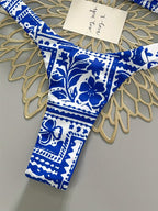 A close-up of a bikini bottom with a bold blue and white floral and geometric pattern, laid flat on a gold-colored decorative doily over a gray surface. A small handwritten note is tucked near the top.