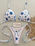 A two-piece bikini set with a white base and blue starburst, eye, and sun patterns, laid flat on a light gray background with decorative props including a small 'Hello' card and a brown woven mat.
