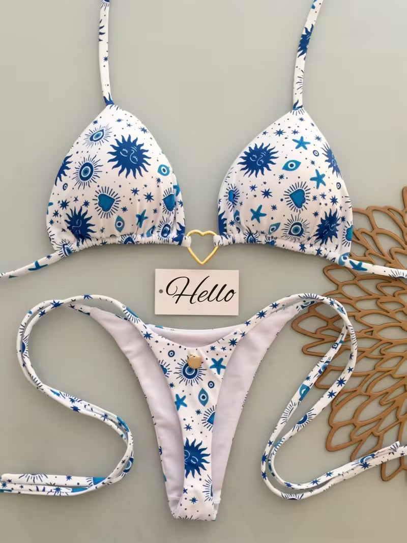 A two-piece bikini set with a white base and blue starburst, eye, and sun patterns, laid flat on a light gray background with decorative props including a small 'Hello' card and a brown woven mat.