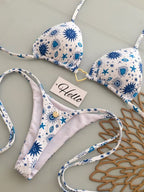 A two-piece triangle bikini set laid flat on a light gray surface. The bikini has a white base with a blue celestial print of stars, suns, and eyes, and features a small gold heart-shaped ring at the center of the top cup.