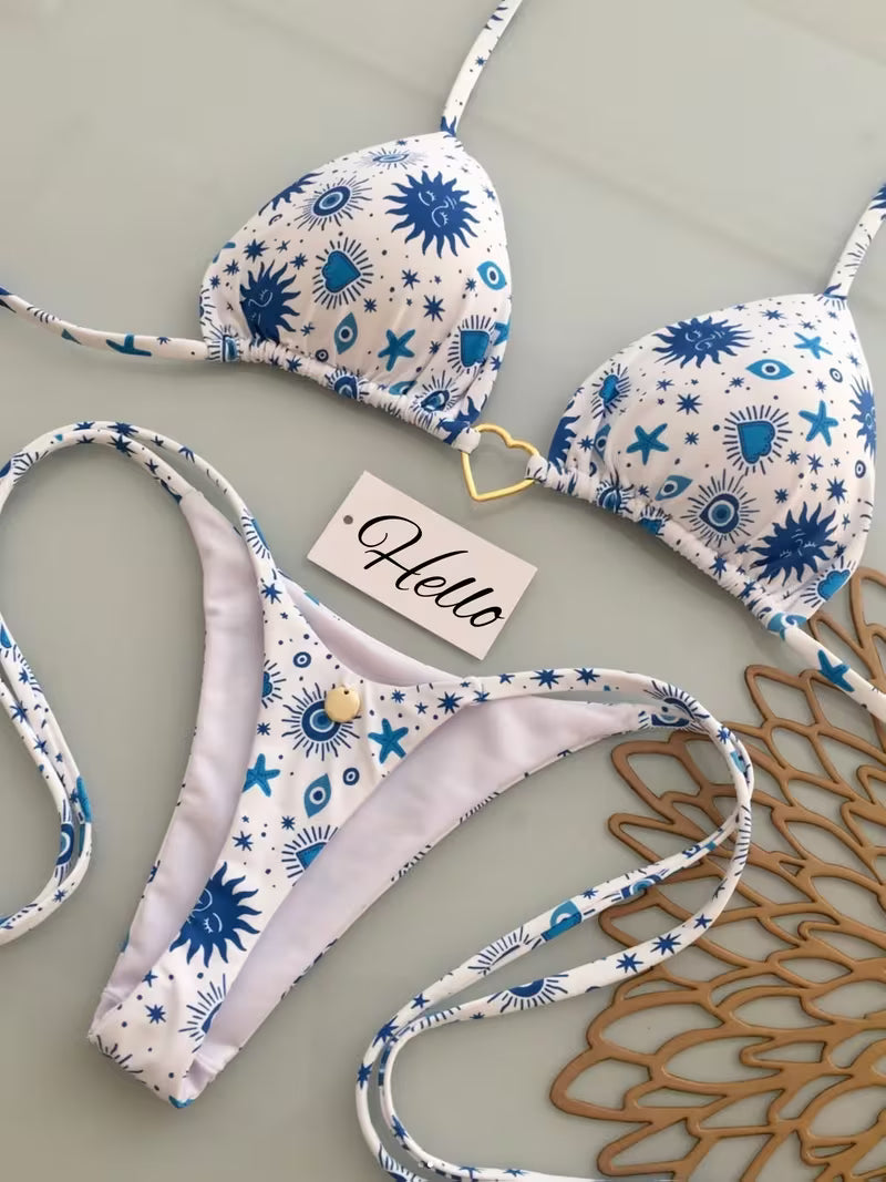 A two-piece triangle bikini set laid flat on a light gray surface. The bikini has a white base with a blue celestial print of stars, suns, and eyes, and features a small gold heart-shaped ring at the center of the top cup.