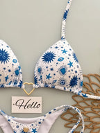 A white string bikini top and bottom set with a blue celestial print of suns, stars, and eyes, styled on a light gray background with a wooden hanger and a small 'Hello' card.
