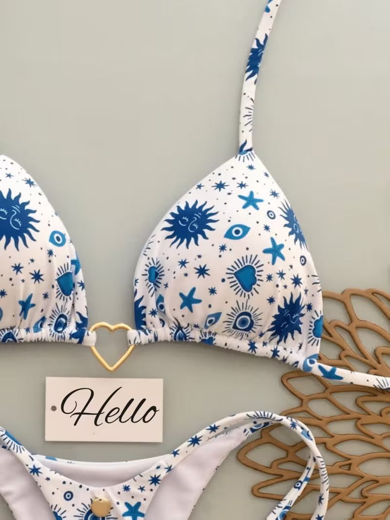 A white string bikini top and bottom set with a blue celestial print of suns, stars, and eyes, styled on a light gray background with a wooden hanger and a small 'Hello' card.