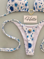 A bikini set laid flat on a light gray surface, featuring a white fabric with blue sun, star, and eye motifs. The top is partially visible at the top of the frame, and the matching thong is spread out below.