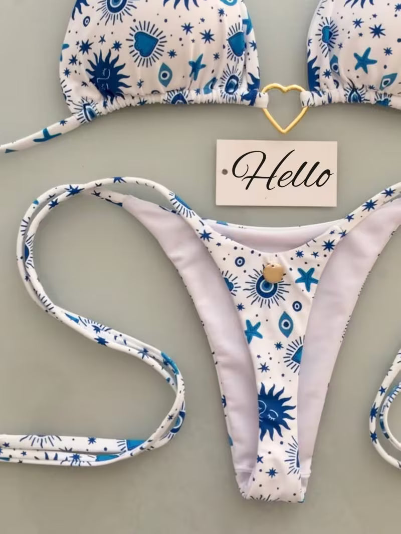 A bikini set laid flat on a light gray surface, featuring a white fabric with blue sun, star, and eye motifs. The top is partially visible at the top of the frame, and the matching thong is spread out below.