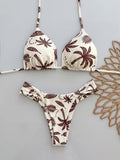 A two-piece triangle bikini set with a brown palm tree and leaf pattern on an off-white background, laid flat on a light gray surface next to a decorative wooden cutout.