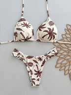 A two-piece triangle bikini set with a brown palm tree and leaf pattern on an off-white background, laid flat on a light gray surface next to a decorative wooden cutout.