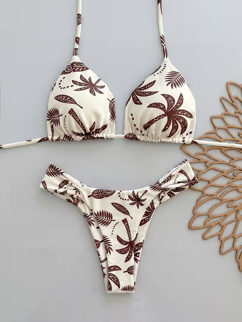 A two-piece triangle bikini set with a brown palm tree and leaf pattern on an off-white background, laid flat on a light gray surface next to a decorative wooden cutout.
