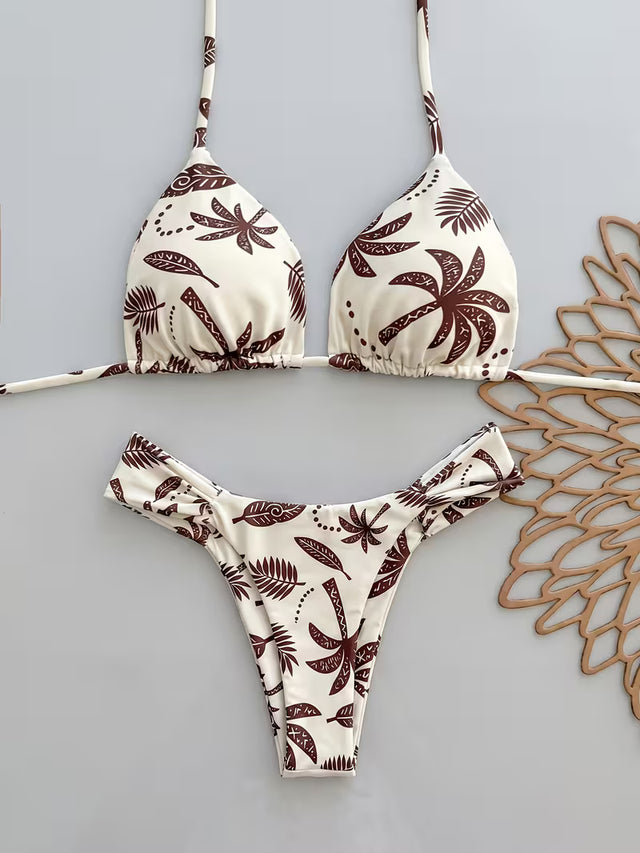A two-piece triangle bikini set with a brown palm tree and leaf pattern on an off-white background, laid flat on a light gray surface next to a decorative wooden cutout.