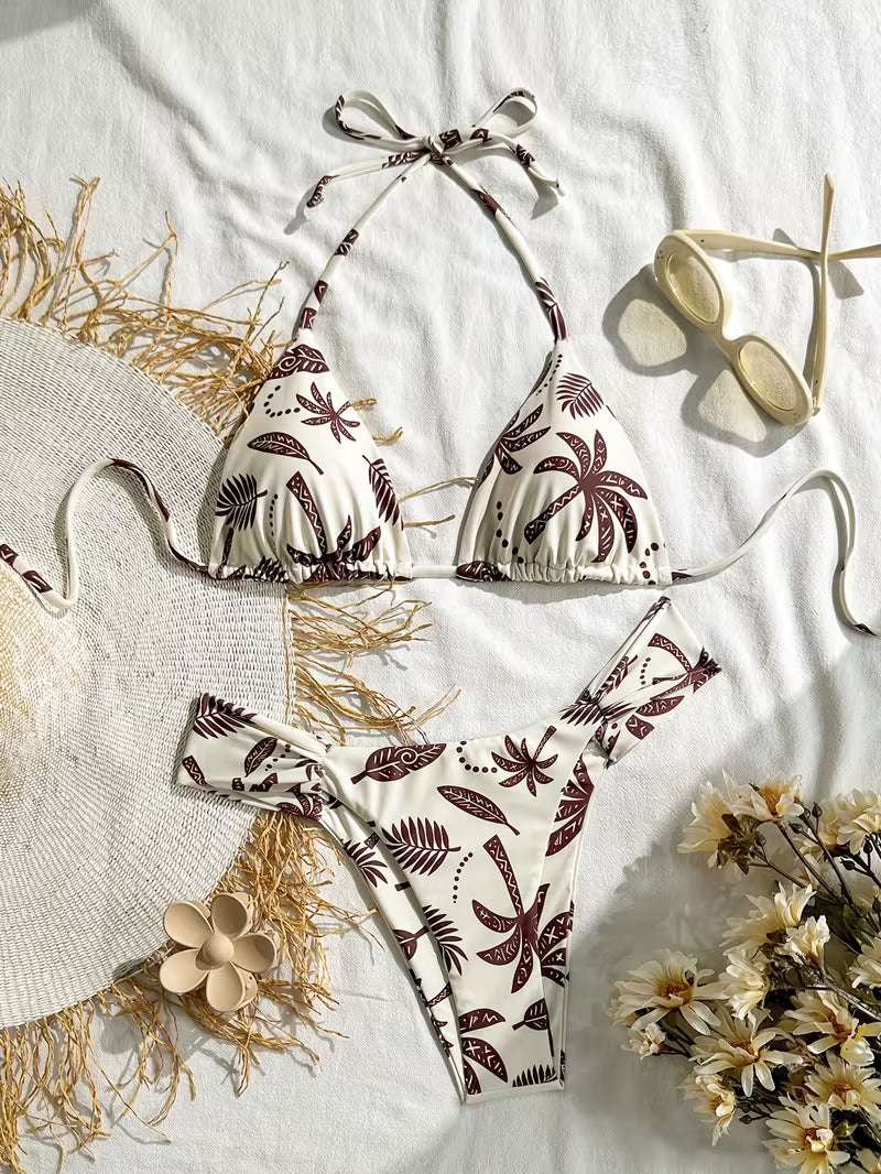 A two-piece string bikini with a brown palm tree and leaf pattern on an off-white background, styled in a flat lay with a straw hat, sunglasses, and flowers on a white fabric backdrop.