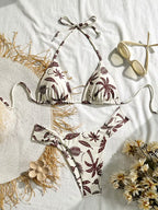 A two-piece string bikini with a brown palm tree and leaf pattern on an off-white background, styled in a flat lay with a straw hat, sunglasses, and flowers on a white fabric backdrop.
