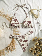 A two-piece bikini set with a brown palm leaf pattern on an off-white background, styled in a flat lay with a straw hat, sunglasses, dried flowers, and a white fabric backdrop.