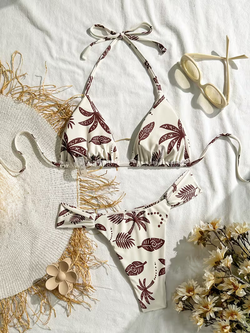 A two-piece bikini set with a brown palm leaf pattern on an off-white background, styled in a flat lay with a straw hat, sunglasses, dried flowers, and a white fabric backdrop.