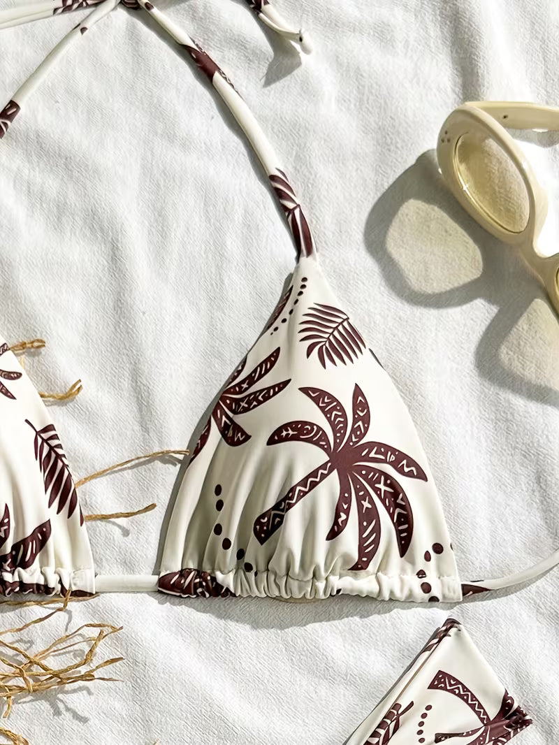 A triangle bikini top laid flat on a white textured fabric background, featuring a cream base with a dark brown palm leaf pattern. A matching bottom is partially visible at the bottom edge, and a pair of beige sunglasses rests in the upper right corner.