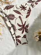 A high-cut bikini bottom with a cream-colored base and a dark brown tribal-style leaf pattern, laid flat on a textured white fabric with dried grass and a flower prop.