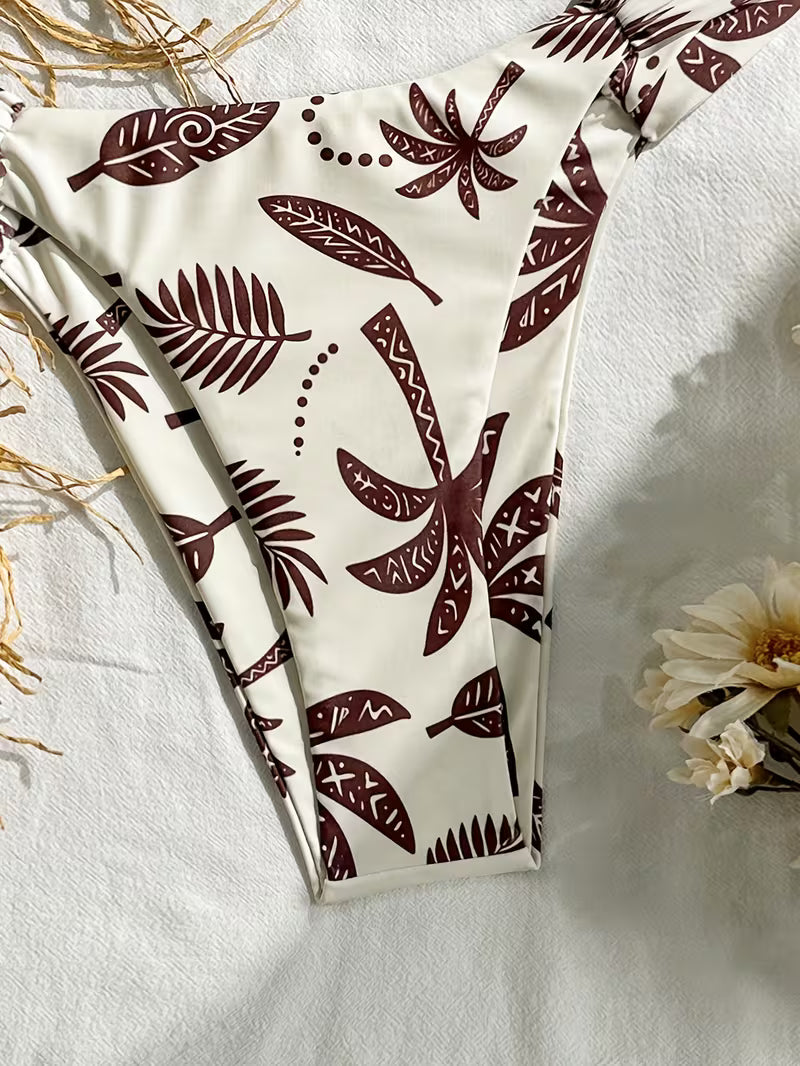 A high-cut bikini bottom with a cream-colored base and a dark brown tribal-style leaf pattern, laid flat on a textured white fabric with dried grass and a flower prop.