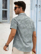A man wearing a light gray short-sleeve button-up shirt with a subtle tropical leaf pattern, shown from the back. He is wearing beige pants, sunglasses, and a bracelet, standing in front of a white wall with blue wooden doors.
