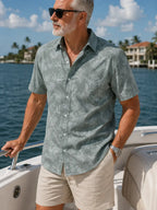 A man wearing a light sage green short-sleeve button-up shirt with a tropical leaf pattern, standing on a boat with water and houses in the background.