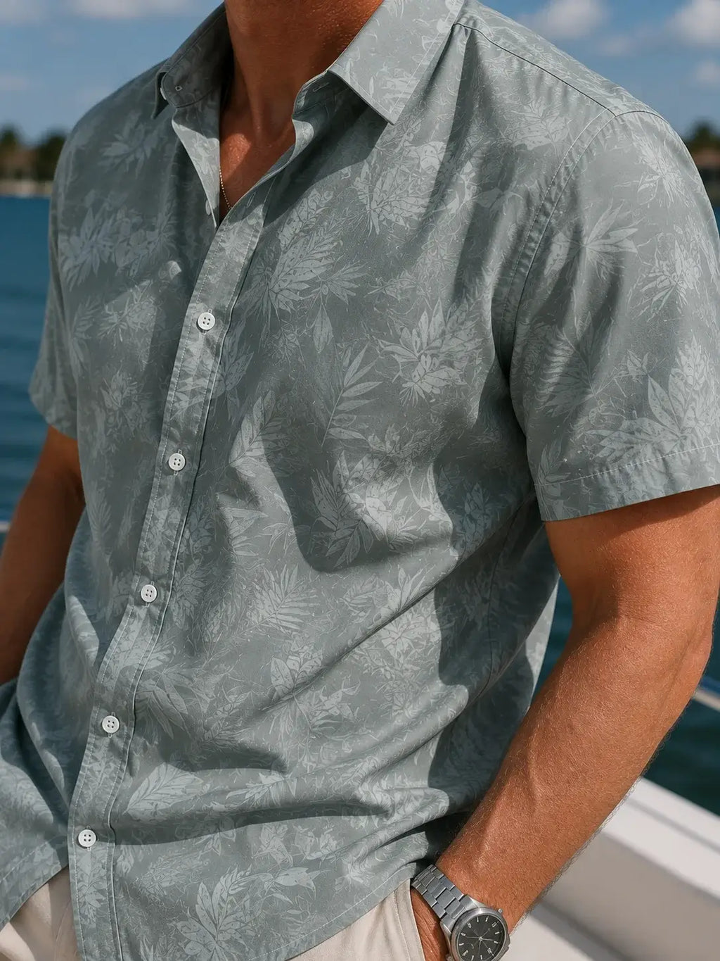 A man wearing a light sage green short-sleeve button-up shirt with a subtle tropical leaf pattern, photographed outdoors near the water.