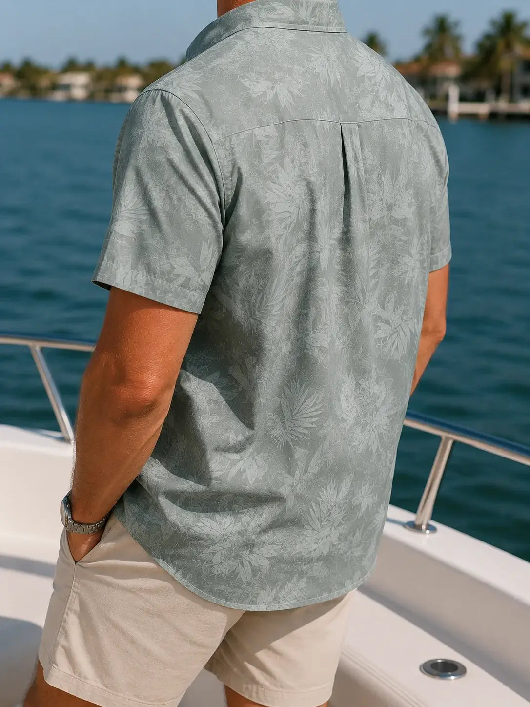 A man wearing a light sage green short-sleeve button-up shirt with a subtle tropical leaf pattern, standing on a white boat deck with blue water and palm trees in the background.