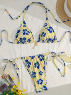 A two-piece string bikini laid flat on a white surface, featuring a pale yellow base with a bold blue floral print. The set includes a triangle top with halter-style ties and matching side-tie bottoms.
