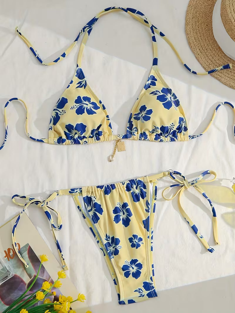A two-piece string bikini laid flat on a white surface, featuring a pale yellow base with a bold blue floral print. The set includes a triangle top with halter-style ties and matching side-tie bottoms.