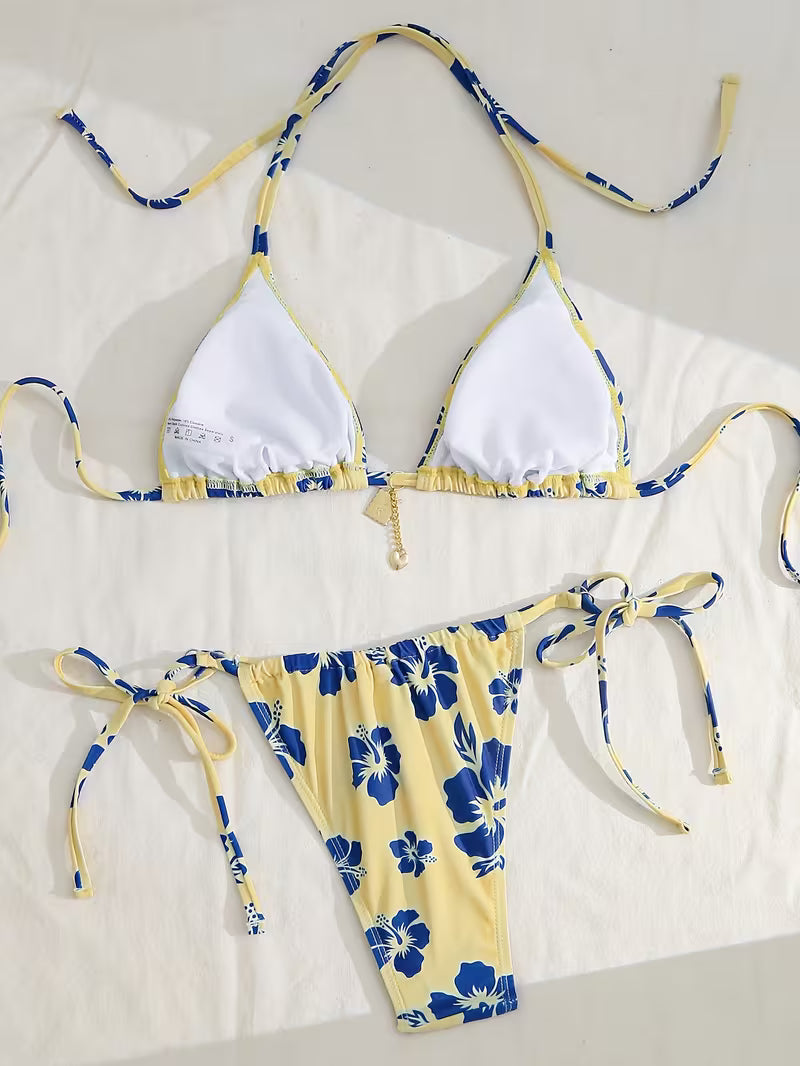 A two-piece string bikini laid flat on a white fabric background. The triangle top has white front panels with yellow and blue floral-patterned straps and trim.
