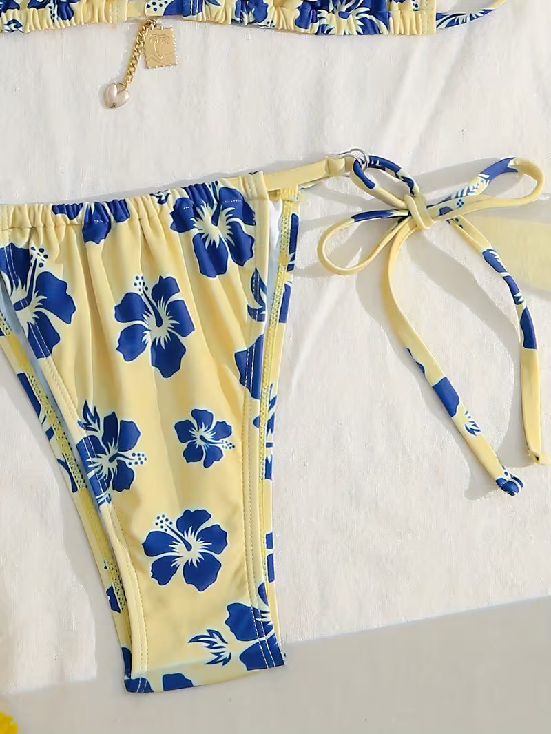 Yellow bikini bottom with a blue hibiscus floral print and tie-side waistband, laid flat on a white background with part of a matching top visible at the top edge.