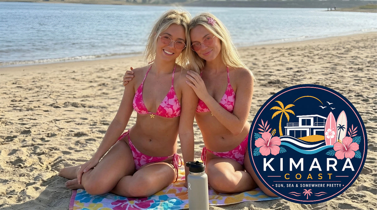 Two women sitting on a beach towel on a sandy beach wearing matching pink floral bikinis. A circular logo for 'Kimara Coast' is visible on the right side of the image.