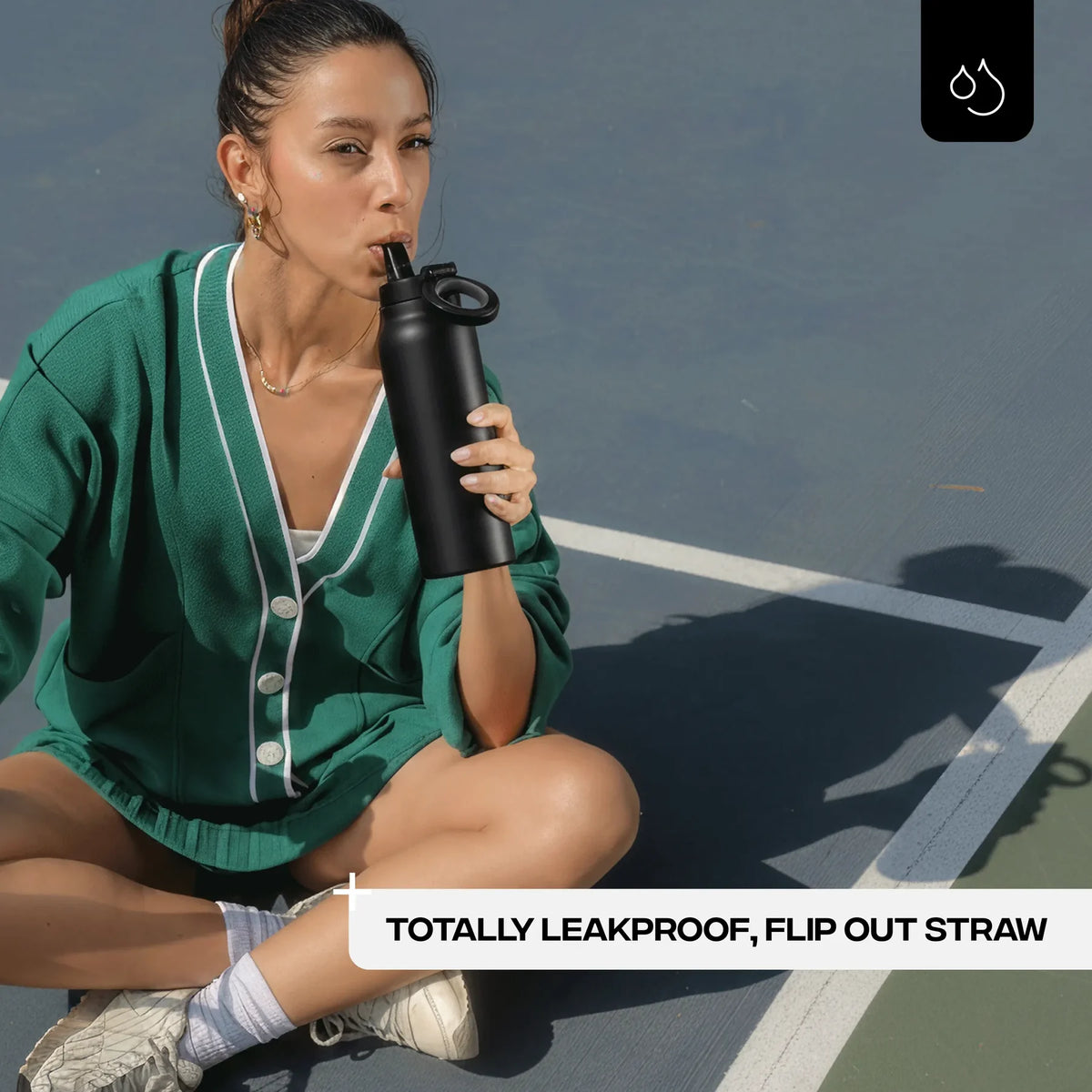 Woman sitting on a tennis court holding a black water bottle with a flip-out straw.