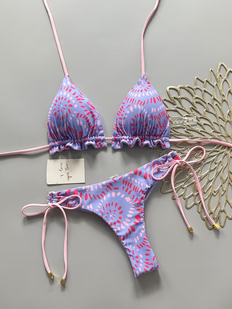 A two-piece bikini set laid flat on a gray background, featuring a light purple and pink abstract pattern with pink strings. The set includes a triangle top with ruffled edges and matching low-rise side-tie bottoms.
