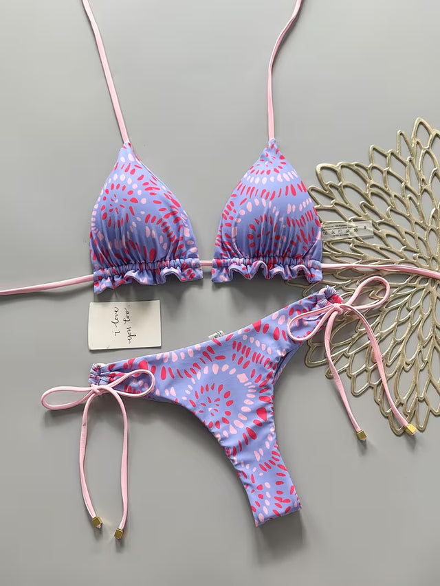 A two-piece bikini set laid flat on a gray background, featuring a light purple and pink abstract pattern with pink strings. The set includes a triangle top with ruffled edges and matching low-rise side-tie bottoms.