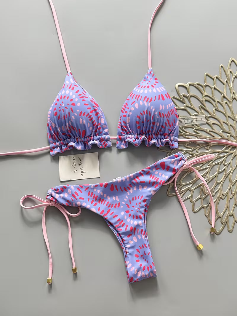 A two-piece bikini set with a light purple and pink abstract pattern, featuring thin pink straps and side-tie bottoms, laid flat on a gray background next to a decorative gold leaf.