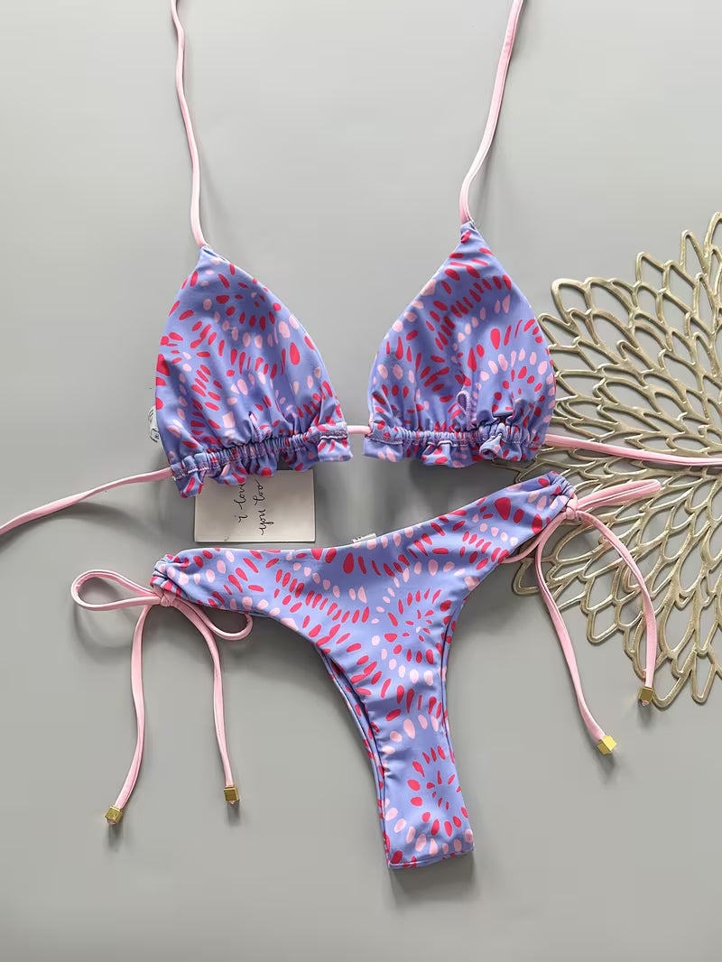 A two-piece string bikini with a lavender and pink abstract pattern, laid flat on a gray background next to a decorative gold fan.