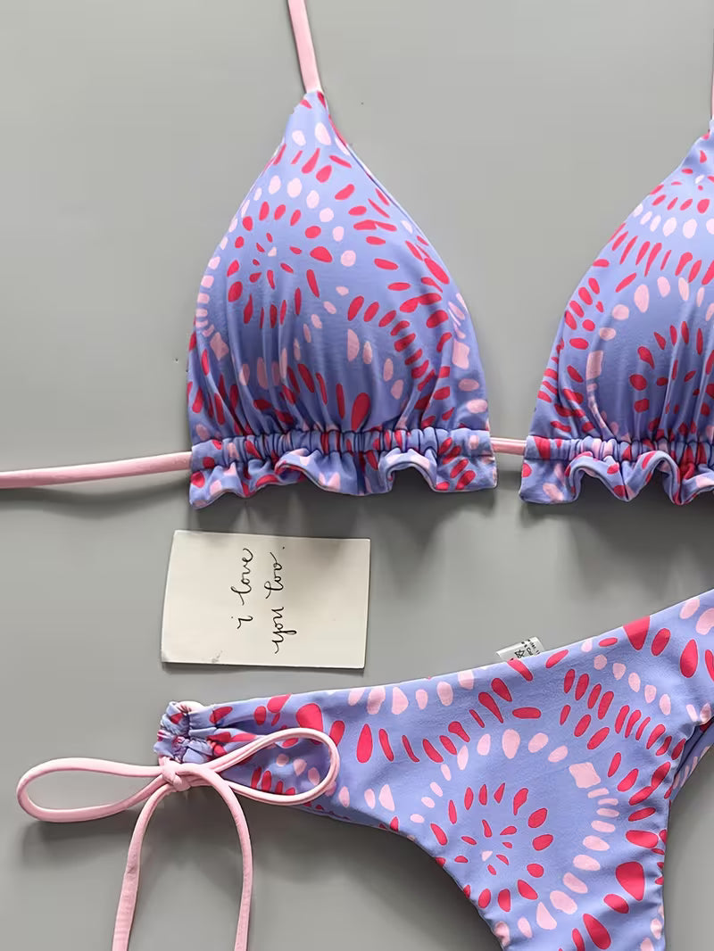 A two-piece string bikini laid flat on a gray background. The triangle top and matching bottoms feature a lavender-blue base with a pink and red abstract spotted pattern, and the thin ties are light pink.