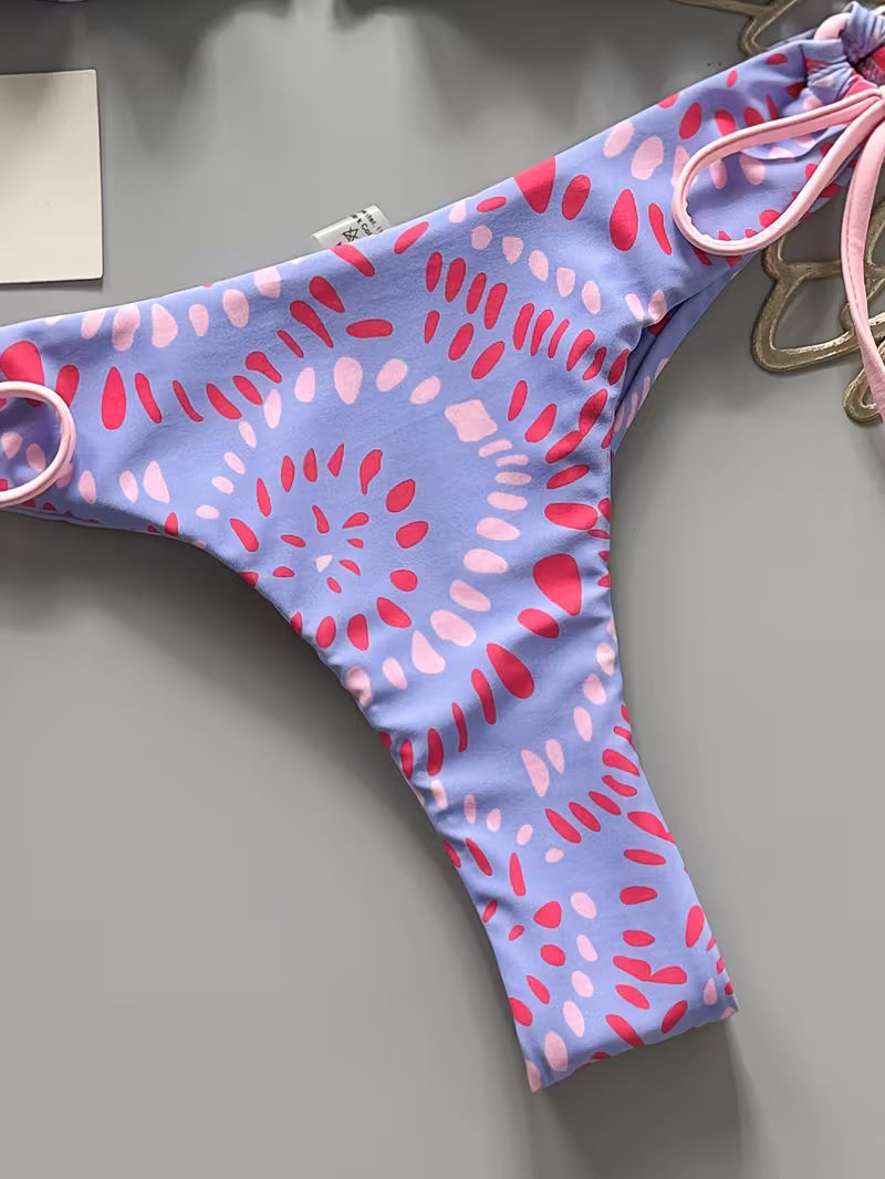 A close-up of a bikini bottom with a light purple base and a pink and red abstract spiral pattern, featuring side ties and a thong cut.