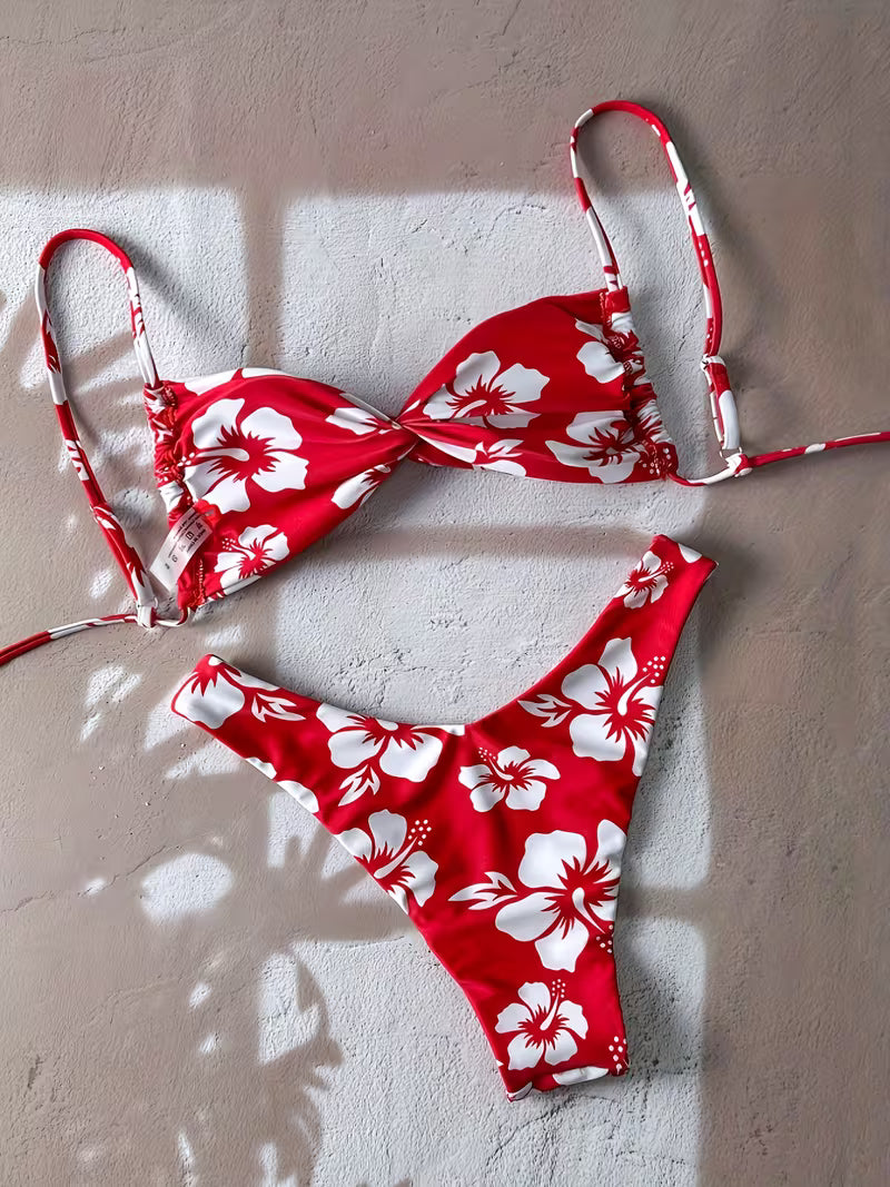 Red and white floral bikini set laid flat on a textured beige wall with soft window shadows. The set includes a triangle bikini top and matching high-cut bikini bottom with a bold hibiscus flower print.
