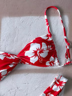 Red and white floral bikini top laid flat on a textured light gray surface, with matching bikini bottoms partially visible at the bottom right.