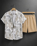 A men's two-piece casual set featuring a white short-sleeve button-up shirt with a gray and beige tropical leaf print, styled over a white undershirt, paired with beige drawstring shorts, hanging on wooden hangers against a dark gray wall.