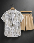 A men's two-piece casual set featuring a white short-sleeve button-up shirt with a gray and beige tropical leaf print, styled over a white undershirt, paired with beige drawstring shorts, hanging on wooden hangers against a dark gray wall.