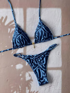 A two-piece bikini set with a blue and white abstract wavy pattern, laid flat on a textured beige wall with soft window shadow overlays.