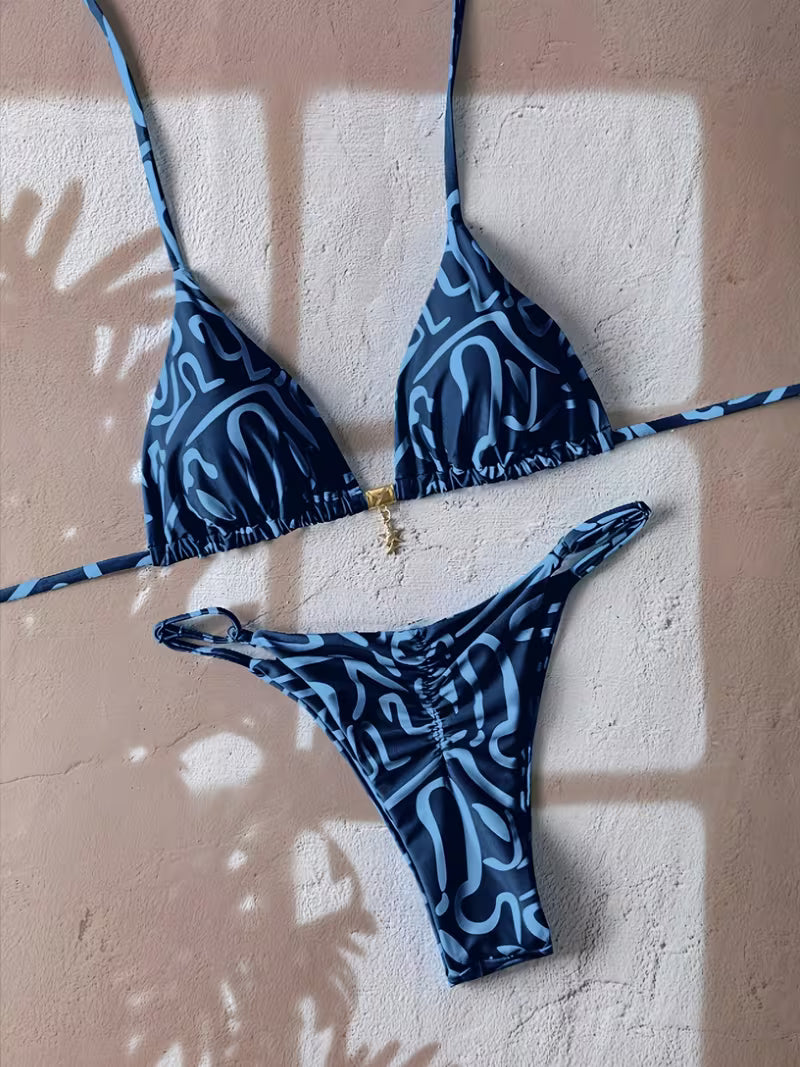 A two-piece bikini set with a blue and white abstract wavy pattern, laid flat on a textured beige wall with soft window shadow overlays.