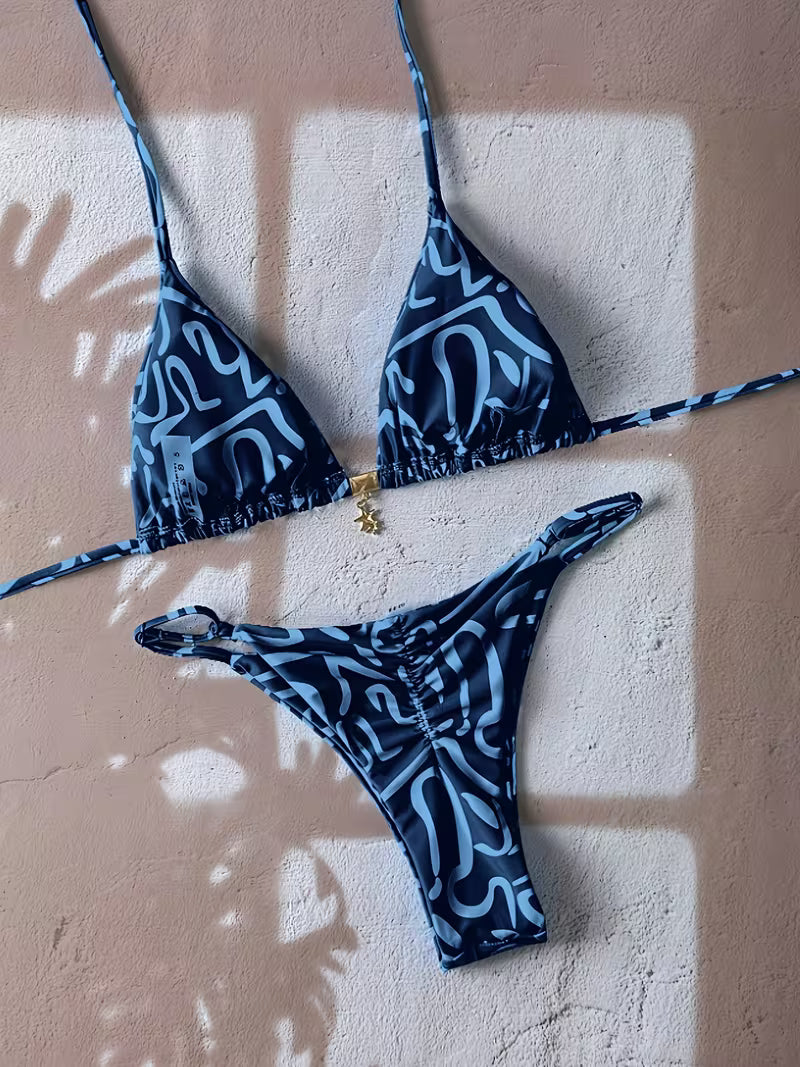 A two-piece bikini set with a dark blue and light blue abstract wavy pattern, laid flat on a textured beige wall with leaf shadows cast across it.