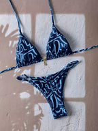 A two-piece bikini set with a dark blue and light blue abstract wavy pattern, laid flat on a textured beige wall with leaf shadows cast across it.