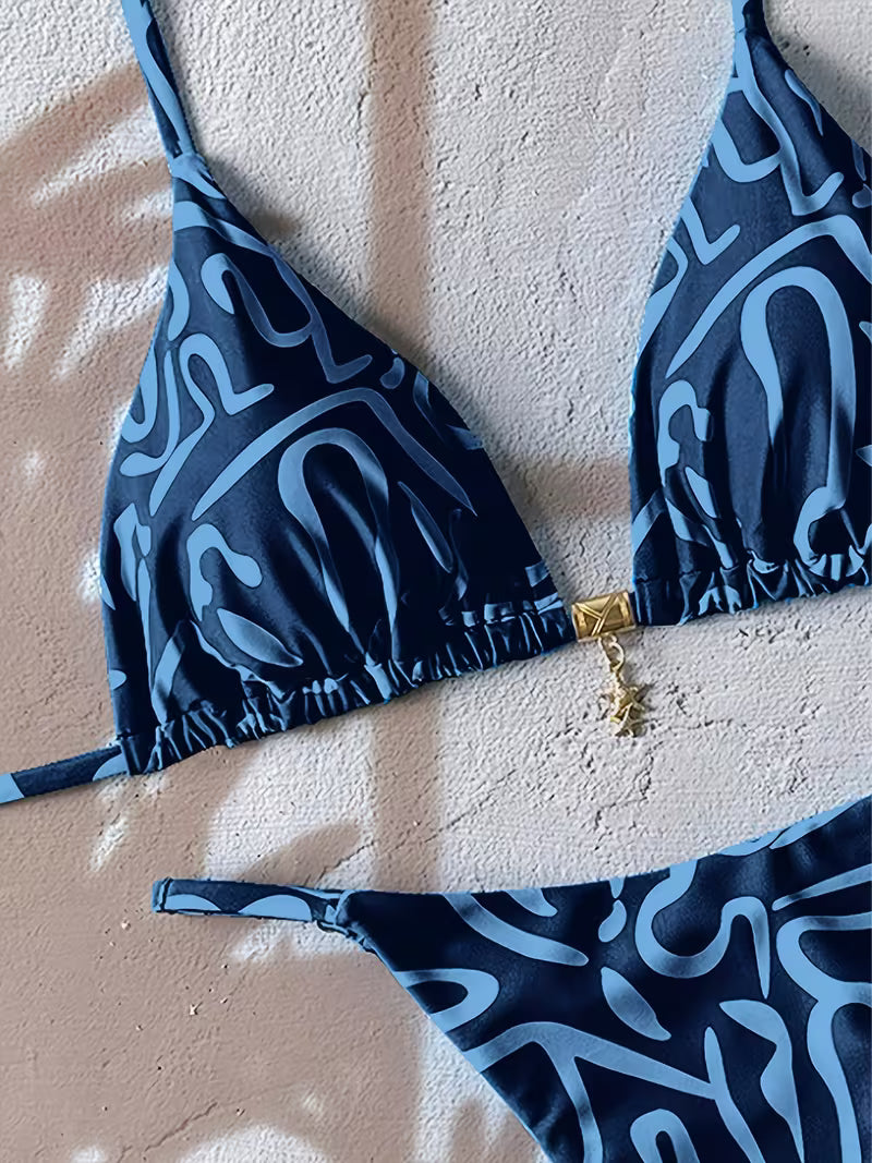 A two-piece bikini set with a dark navy blue and light blue abstract wavy pattern, featuring a triangle top with a gold zipper detail and a matching low-rise bottom, laid flat on a textured beige wall with leaf shadows.