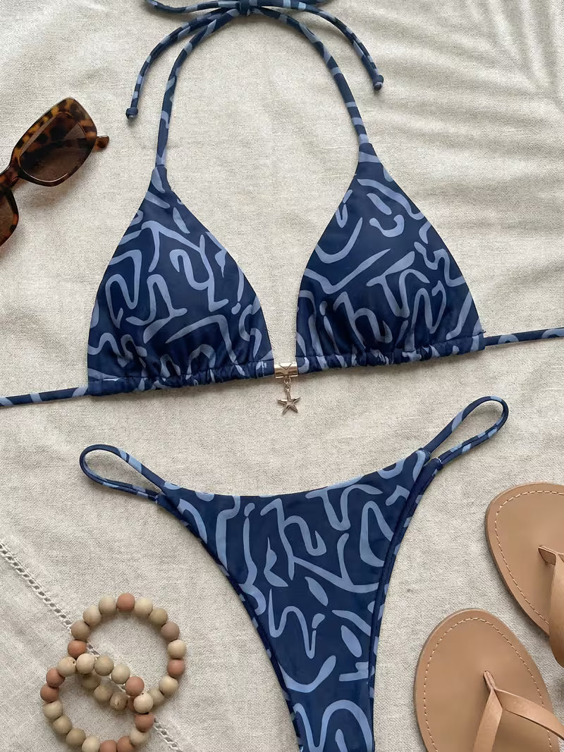 A two-piece string bikini with a blue and light blue abstract wavy pattern, laid flat on a beige textured blanket alongside sunglasses, a beaded bracelet, and tan flip-flops.