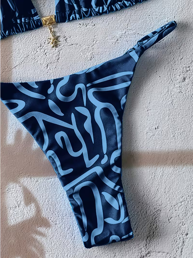 A close-up of a bikini bottom in dark navy blue with a light blue abstract line pattern, laid flat on a textured beige surface.