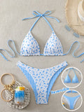 A two-piece bikini set laid flat on a light beige textured background, featuring a white base with blue floral print on one side and blue gingham check on the other. The set includes a halter triangle bikini top and matching low-rise bikini bottoms.